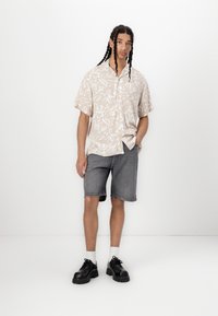 Kortermet beige skjorte med knapper og hvit abstrakt mønster, kombinert med grå denimshorts og svarte sko med kraftig såle. Hvite sokker synlige.