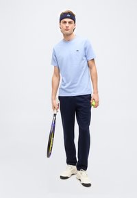 Νέος άνδρας με ανοιχτό μπλε μπλουζάκι, σκούρα μπλε παντελόνια, λευκά αθλητικά παπούτσια και σκούρο μπλε κορδέλα στο κεφάλι, κρατώντας ρακέτα και μπάλα του τένις, στέκεται μπροστά σε απλό φόντο.