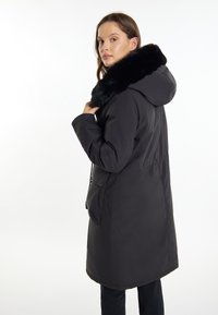 Parka nera con colletto in finitura pelliccia sintetica, cappuccio, tasche frontali, vita aderente e texture liscia, mostrata da dietro.