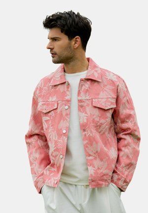 Homme portant une veste rose à motif floral sur une chemise blanche et un pantalon blanc, regardant vers sa gauche, les mains dans les poches.