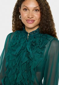 Blouse en chiffon teal avec un devant à volants et un col montant. Détail avec des boutons et une texture douce et transparente, créant un effet superposé.