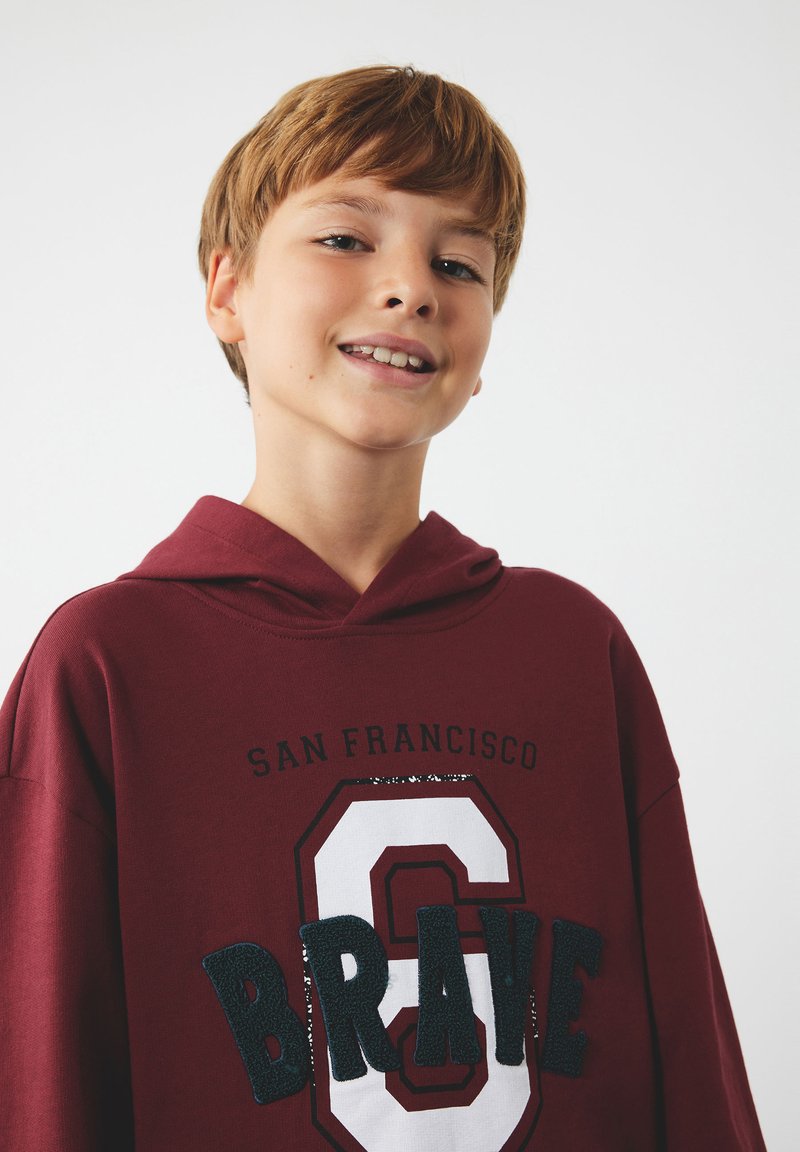 Bordowy hoodie z dużą grafiką z napisem "BRAVE" w teksturowanym granacie oraz białym heksagonalnym kształtem. Posiada kaptur ze sznurkiem i luźny krój.