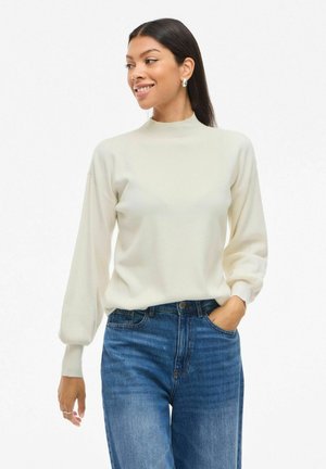 Pull blanc à col montant avec des manches ballon, présentant une texture douce, associé à un jean taille haute bleu foncé.