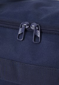 Zoom sur un sac en tissu bleu marine doté de doubles tirettes de fermeture éclair avec un logo, une surface texturée et un détail de couture renforcée à la base.