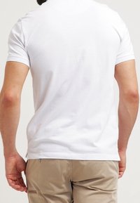 Homme portant un polo blanc uni à manches courtes et un pantalon marron clair, vu de dos avec les mains lâchées sur les côtés.