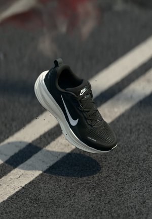 Sort-hvid løbesko i luften over en asfaltvej med hvid linje, der viser "RUN" og Nike swoosh-logoerne.