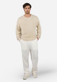 Beige fluffy sweater with a round neck, paired with loose white linen pants featuring large patches. Model wears white sneakers.