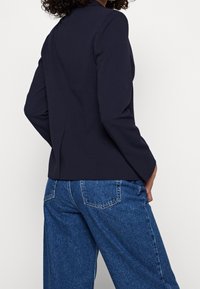 Marinblå kavaj med långa ärmar, skräddarsydd passform och subtil sömdetaljering, kombinerad med högmidjade ljusblå denimbyxor.