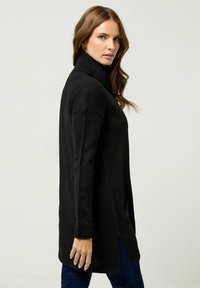 Maglione nero a collo alto con lavorazione a maglia texturizzata, maniche lunghe, spacchi laterali e vestibilità ampia, abbinato a jeans blu scuro.