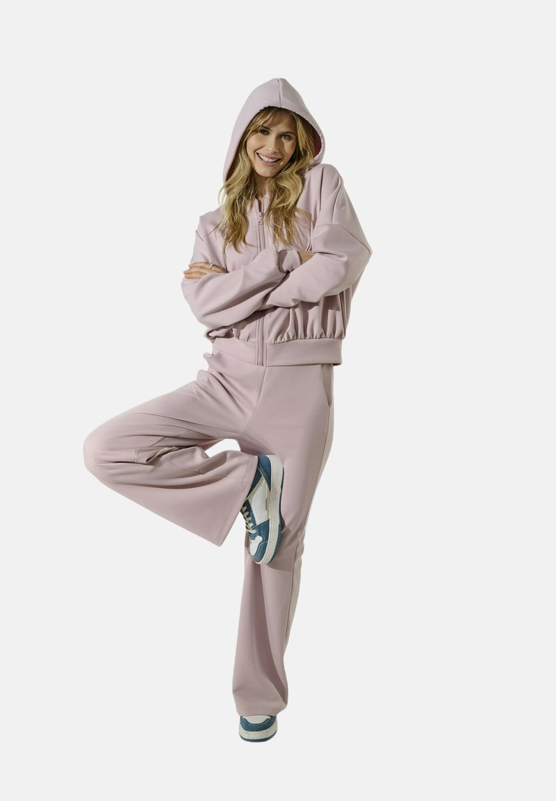 Donna che sorride con le braccia incrociate, indossando una felpa leggera rosa e pantaloni larghi abbinati, in equilibrio su una gamba con le scarpe da ginnastica.