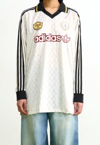 Bílý dlouhý rukáv dres s texturovaným diamantovým vzorem, černým límcem a třemi černými pruhy na rukávech. Obsahuje logo Adidas a odznaky.