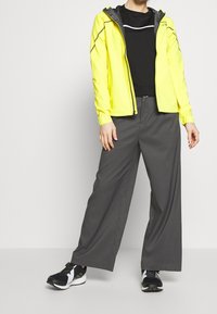 Persona con un giubbotto giallo neon sopra una maglietta nera, pantaloni grigi larghi e sneakers bianco e nere, in piedi contro uno sfondo neutro.