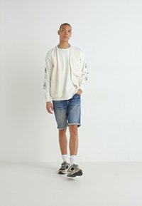Chemise blanche à manches longues avec poches, shorts en denim et baskets. La chemise présente des motifs imprimés noirs sur les manches.