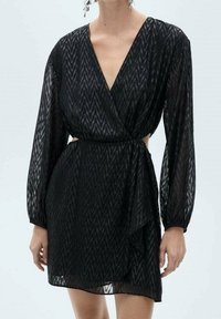 Czarna, długorękawowa mini sukienka w zygzakowy wzór, z głębokim dekoltem w kształcie V, wyciętymi bokami i sukienką w stylu kopertowym na stojącej osobie.