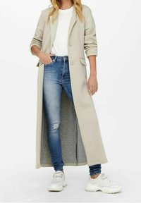 Abrigo largo beige con dos botones y bolsillos laterales, llevado sobre una camisa blanca y vaqueros azules con desgastes. Completado con zapatillas blancas.