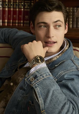 Silver stainless steel watch with a black dial and gold accents; worn on a hand resting on a denim jacket. Background features bookshelves.
