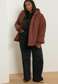 Brązowa kurtka puchowa z kapturem, z pikowanym wzorem i zapięciami na napy, noszona na czarnej bluzce i szerokich czarnych spodniach.