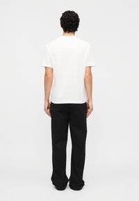 Άνδρας με σγουρά μαλλιά που φοράει ένα απλό λευκό κοντομάνικο μπλουζάκι και φαρδιά μαύρη παντελόνια, στέκεται με την πλάτη προς το μέρος μας σε ένα απλό φωτεινό φόντο.