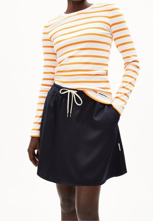Personne portant une chemise blanche à manches longues avec des rayures orange et une jupe noire avec un cordon blanc, main dans la poche de la jupe.