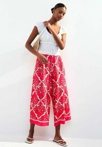 Weit geschnittene Hosen mit einem lebhaften roten Blumenmuster auf weißem Hintergrund, kombiniert mit einem weißen strukturierten Oberteil und schwarzen Sandalen.