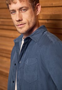 Dark blue corduroy shirt with a buttoned front, collar, and a chest pocket. Features vertical ribbing texture and black buttons.