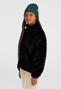 Zwarte pluche fleece jas met hoge kraag en ritsluiting, gedragen over lichtbeige broeken. Het model draagt een teal gebreide beanie met een logo.