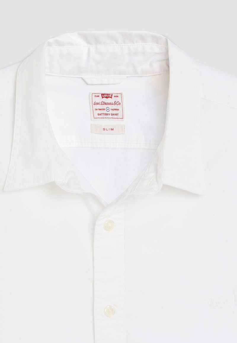 Chemise col montant ajustée blanche en coton, avec une étiquette Levi's portant la mention 'Battery Shirt' et des fermetures à boutons. Texture douce.