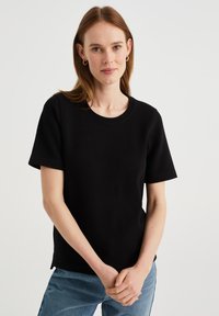 Czarny t-shirt z krótkim rękawem, wykonany z teksturowanej tkaniny, z okrągłym dekoltem i bocznymi rozcięciami, zestawiony z jasnoniebieskimi dżinsami.