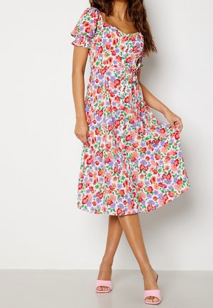 Femme portant une robe midi colorée avec un imprimé floral rouge, violet et rose, des manches bouffantes courtes et des talons ouverts roses.