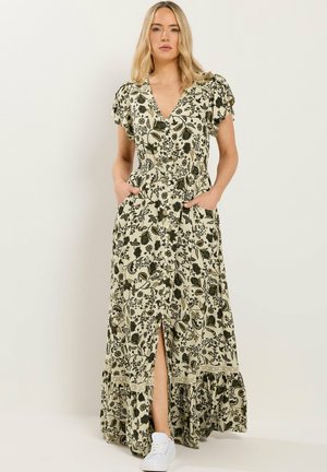 Blonde vrouw staat met handen in haar zakken, draagt een crèmekleurige maxi-jurk met groene bloemenprint en witte sneakers.