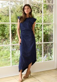 Abito blu navy con maniche a cappuccio, dettagli arricciati, orlo asimmetrico, tessuto testurizzato, abbinato a tacchi beige con cinturini.