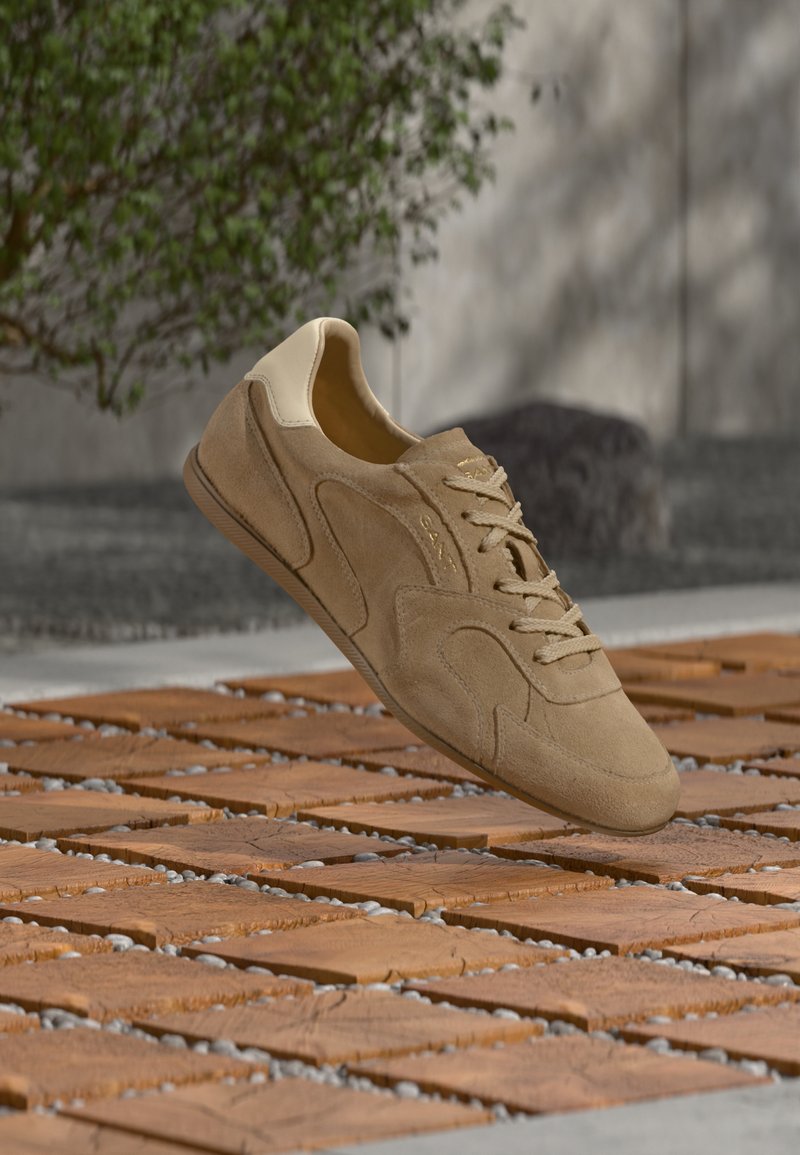 Baskets en daim beige avec talon crème flottant au-dessus d'un sol en carreaux de bois et galets, cadre extérieur avec arbre flou et mur en béton en arrière-plan.