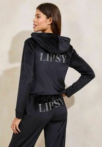 Crni trenirka s kapuljačom, izrađena od baršunaste tkanine. "LIPSY" logo s kamenčićima na stražnjoj strani i hlačama, s dugim rukavima i ležernim krojem.