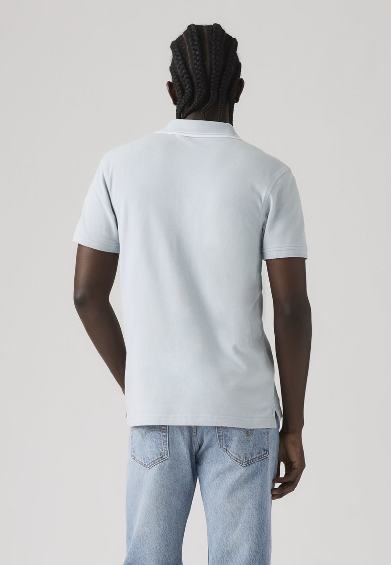 Hombre con cabello trenzado, vistiendo una camiseta polo de manga corta azul claro y vaqueros azul claro, de espaldas sobre un fondo sencillo.