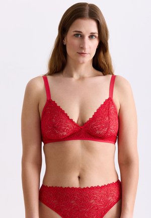 Frau mit heller Haut und langen braunen Haaren trägt passend rotes Spitzen-BH und Slip vor einem einfarbigen weißen Hintergrund.