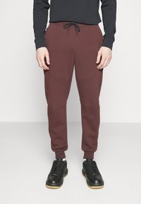 Bordové tepláky s elastickým pasem a šňůrkou, s bočními kapsami a tapered nohavicemi, v kombinaci s černými sportovními botami.