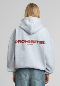 Felpa grigia con una grande scritta rossa "PROIBITO" e "10119, BERLINO" sottostante. Presenta un cappuccio ampio e una vestibilità morbida.