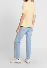 Žluté tričko s krátkým rukávem a kulatým výstřihem, nošeno s světlými modrými džíny rovného střihu a bílými vysokými teniskami s logem značky.