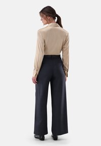 Beige Satinbluse mit langen Ärmeln und Manschetten, kombiniert mit dunkelgrauen Hosen mit weitem Bein. Glatte Textur, maßgeschneiderte Passform und dezente Musterdetails.