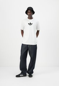 Witte katoenen t-shirt met een zwart Adidas-logo, gecombineerd met donkerblauwe denim jeans en zwarte sneakers met witte strepen. Zwarte bucket hat.