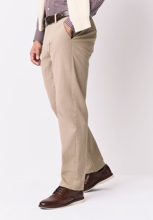 Muž nosiaci béžové nohavice, hnedý kožený opasok, kockovanú košeľu, krémový sveter prehodený cez ramená a tmavohnedé kožené topánky.