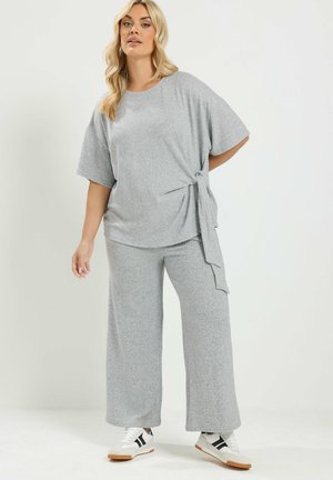 Vrouw met lang blond haar, gekleed in een grijs kortemouwtop met zijstrik en bijpassende wijde broek, staand met één hand achter haar rug.