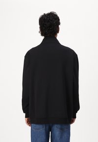 Black pullover jacket with a collar and ribbed cuffs, featuring a smooth texture and relaxed fit, paired with blue denim jeans.