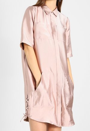 Femme portant une robe chemise à manches courtes rose pâle brillante avec des poches à l'avant et un détail lacé sur le côté.