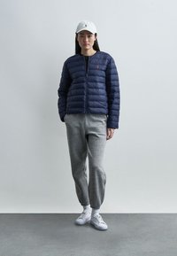 Γυναίκα που φοράει μπλε σκούρο μπουφάν, γκρι φόρμα, λευκά αθλητικά παπούτσια και λευκό καπέλο, στεκόμενη μπροστά σε έναν μονόχρωμο γκρι τοίχο.