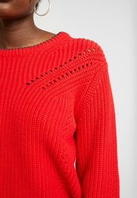 Rød strikket sweater med rund halsudskæring, tekstureret stof og detaljerede åbne masker langs skuldrene. Blødt materiale, lange ærmer.