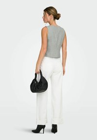 Femme portant un haut gris sans manches, un pantalon large blanc et des bottines à talons noires, tenant un sac à main noir à ses côtés.