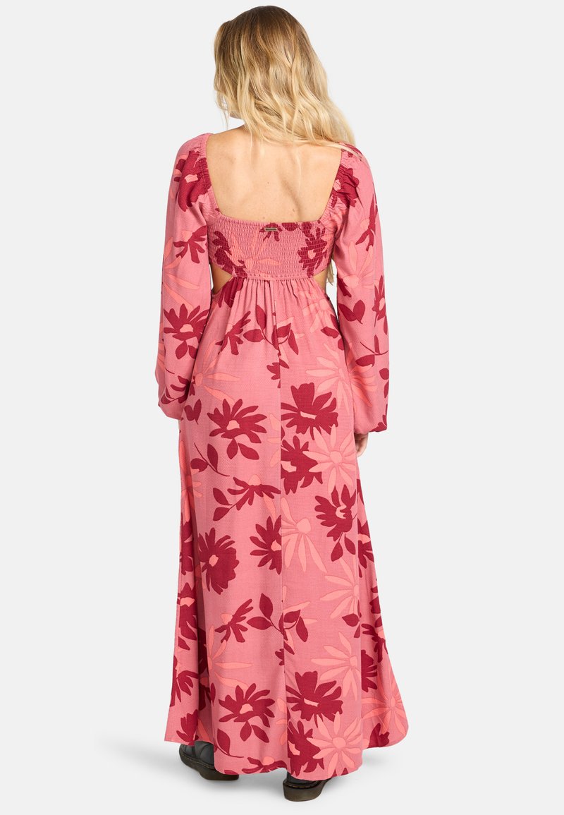 Donna con lunghi capelli biondi, che indossa un maxi abito floreale rosa con maniche lunghe e schiena scoperta, in piedi su uno sfondo bianco.