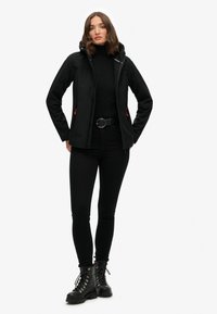 Zwarte softshell jas met een rits aan de voorkant, verstelbare capuchon en rode accenten; gecombineerd met een zwarte turtleneck, skinny jeans en veterschoenen.