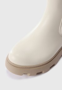 Botte de cheville en caoutchouc beige avec une finition lisse et brillante, semelle texturée tan en contraste, et bout rond. Détails latéraux avec un accent de motif.